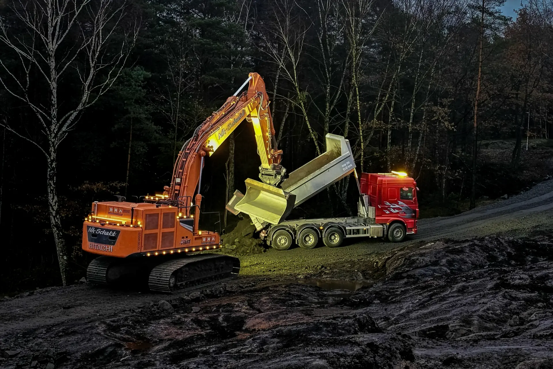 Orange grävmaskin lastar röd lastbil i skymning
